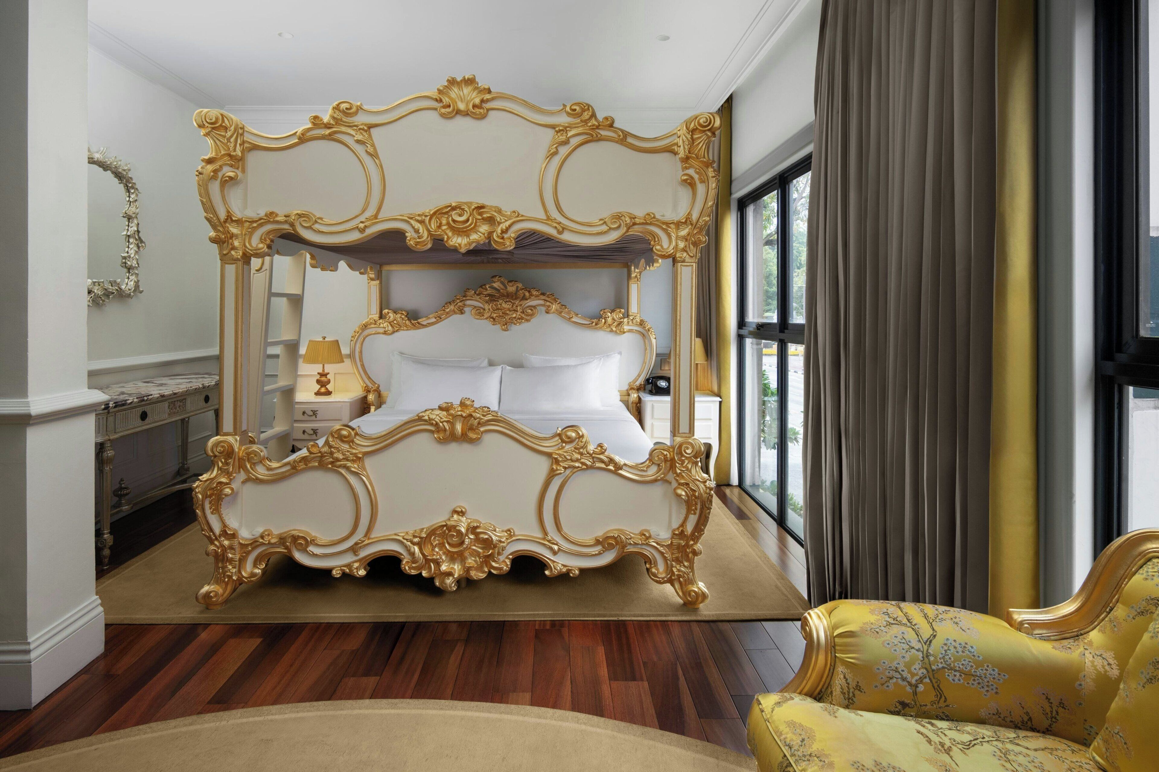 A luxurious bedroom with an ornate four-poster bed, a nightstand, a lamp, and a view of the outdoors through a large window.