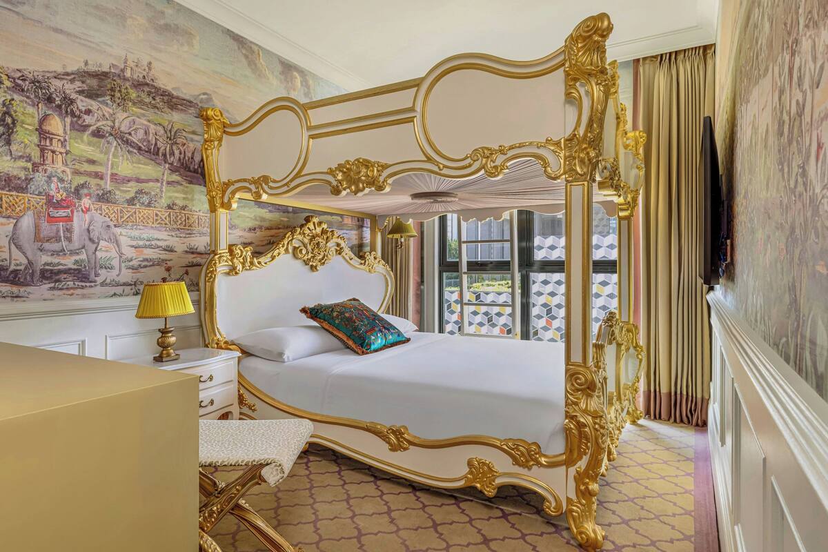 Un lussuoso letto a baldacchino con decorazioni dorate elaborate, un grande affresco sulla parete e vista su un giardino decorato attraverso una finestra.