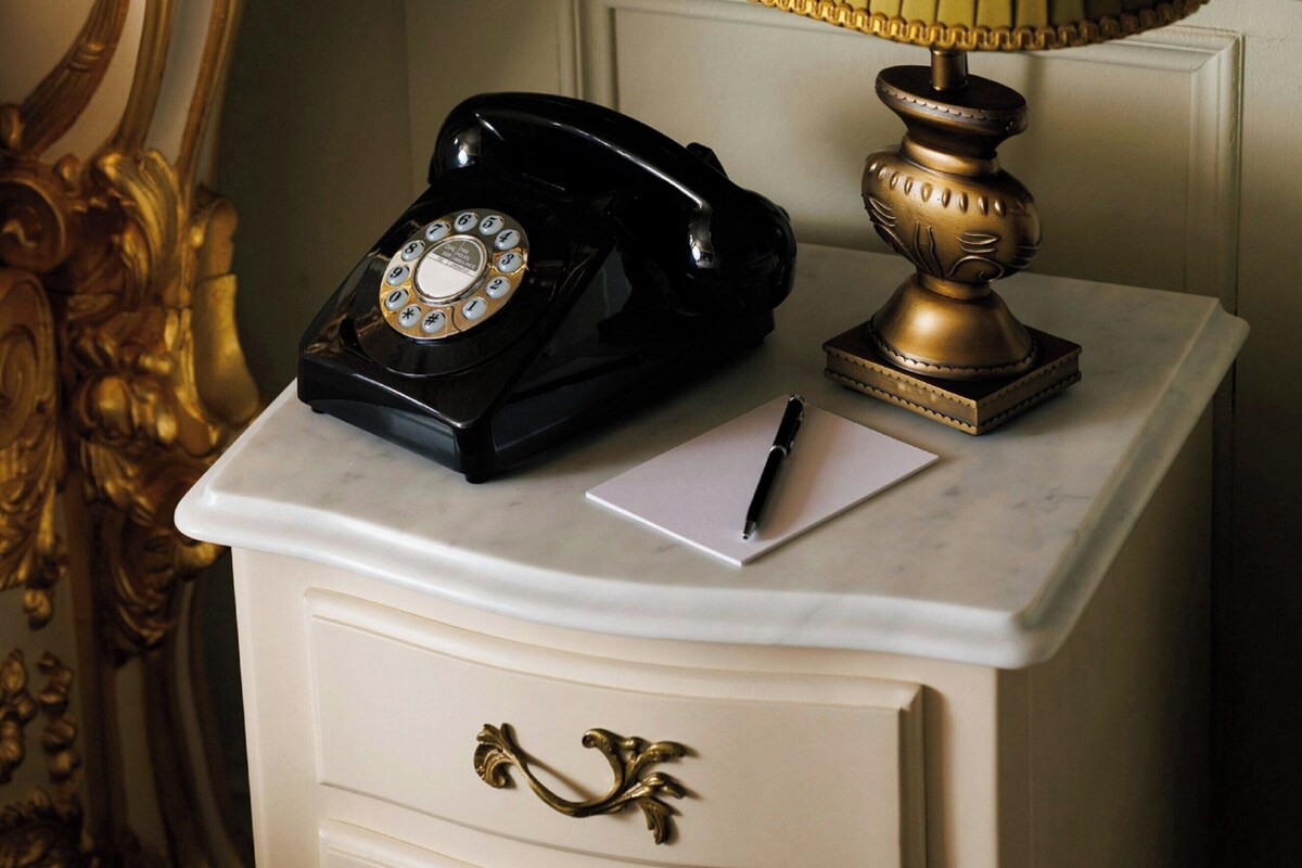 Een oude telefoon op een witte nachtkastje, samen met een lamp, notitieboekje en pen.