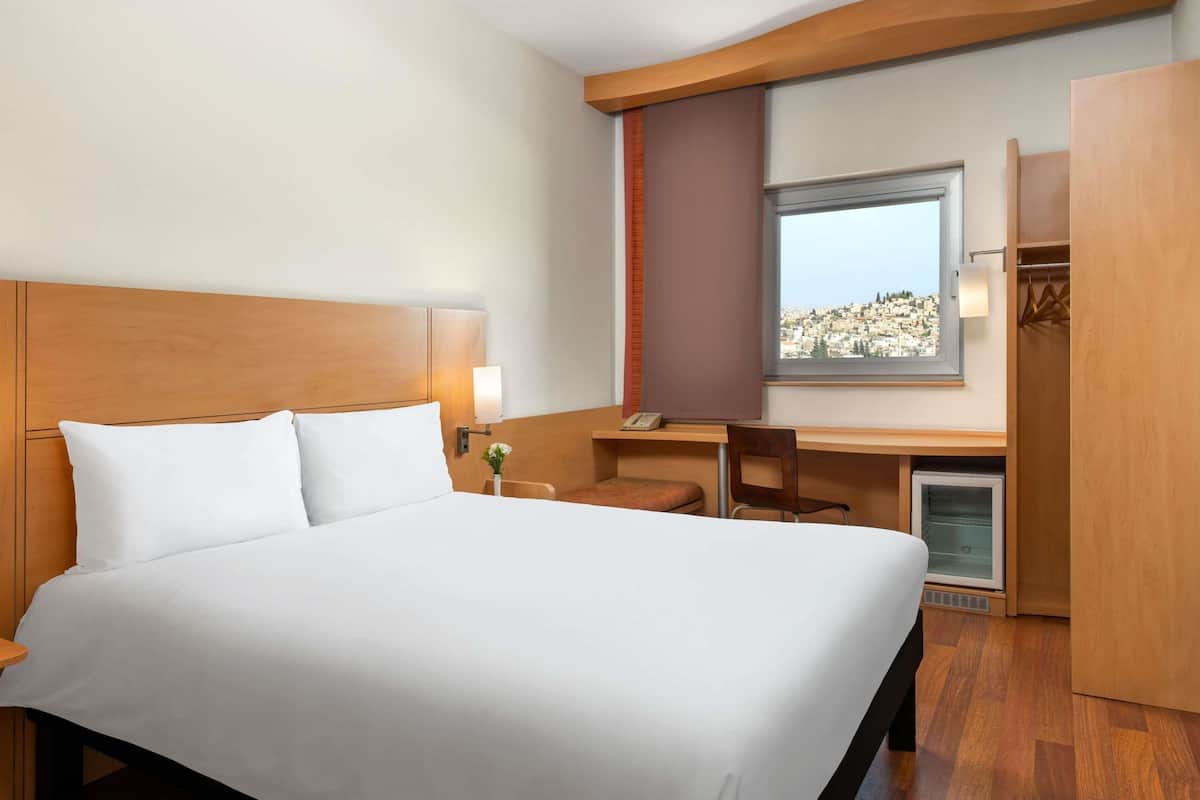 Habitación de hotel con una cama grande, un escritorio con silla, un refrigerador pequeño y una ventana con vista a la ciudad.