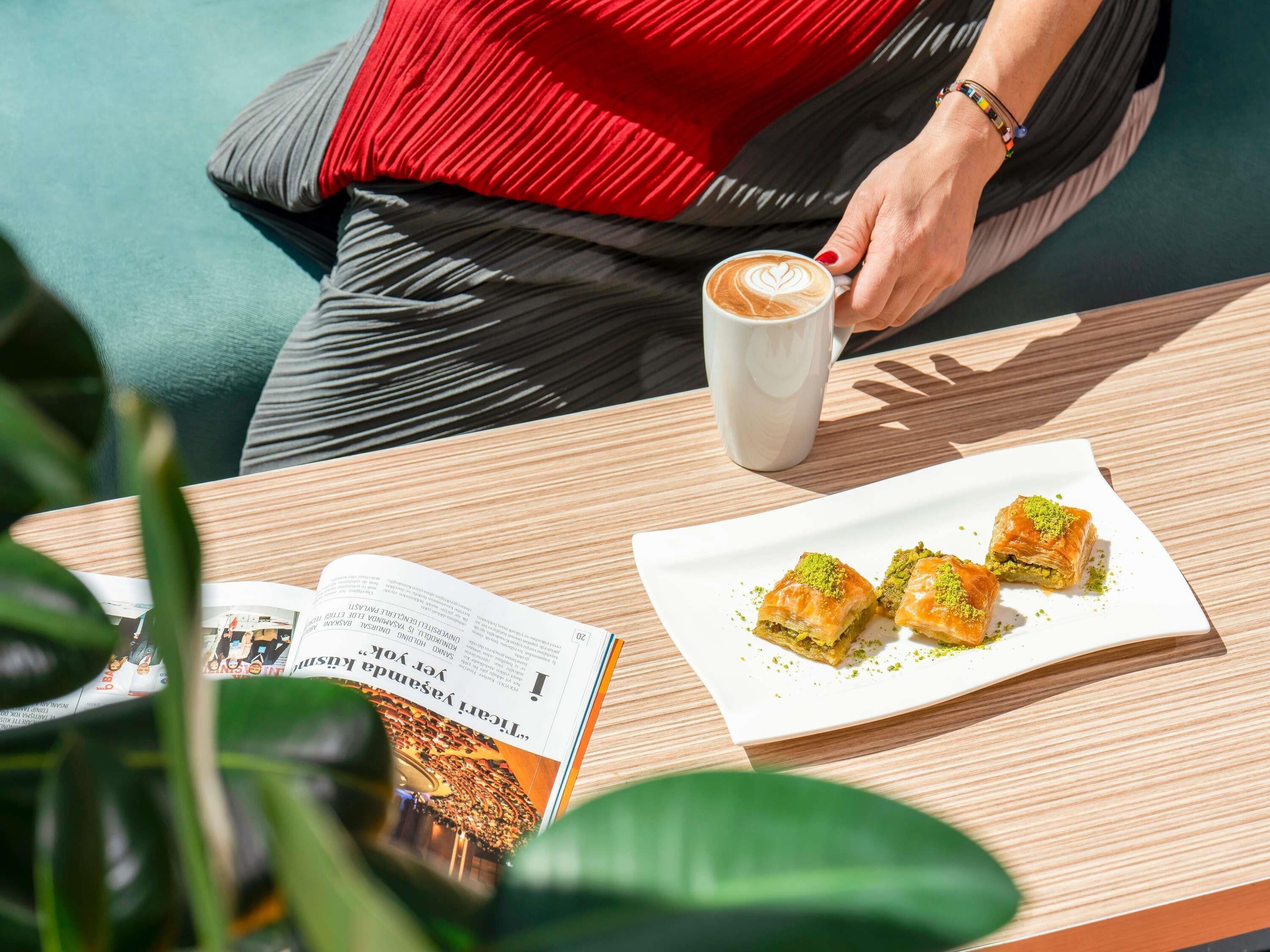 Une personne assise à une table, devant une tasse de café, une assiette de baklava et un magazine.