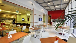 A modern restaurant with white chairs and wooden tables, a red accent wall, and a television screen. - ibis Gaziantep (Gaziantep)
