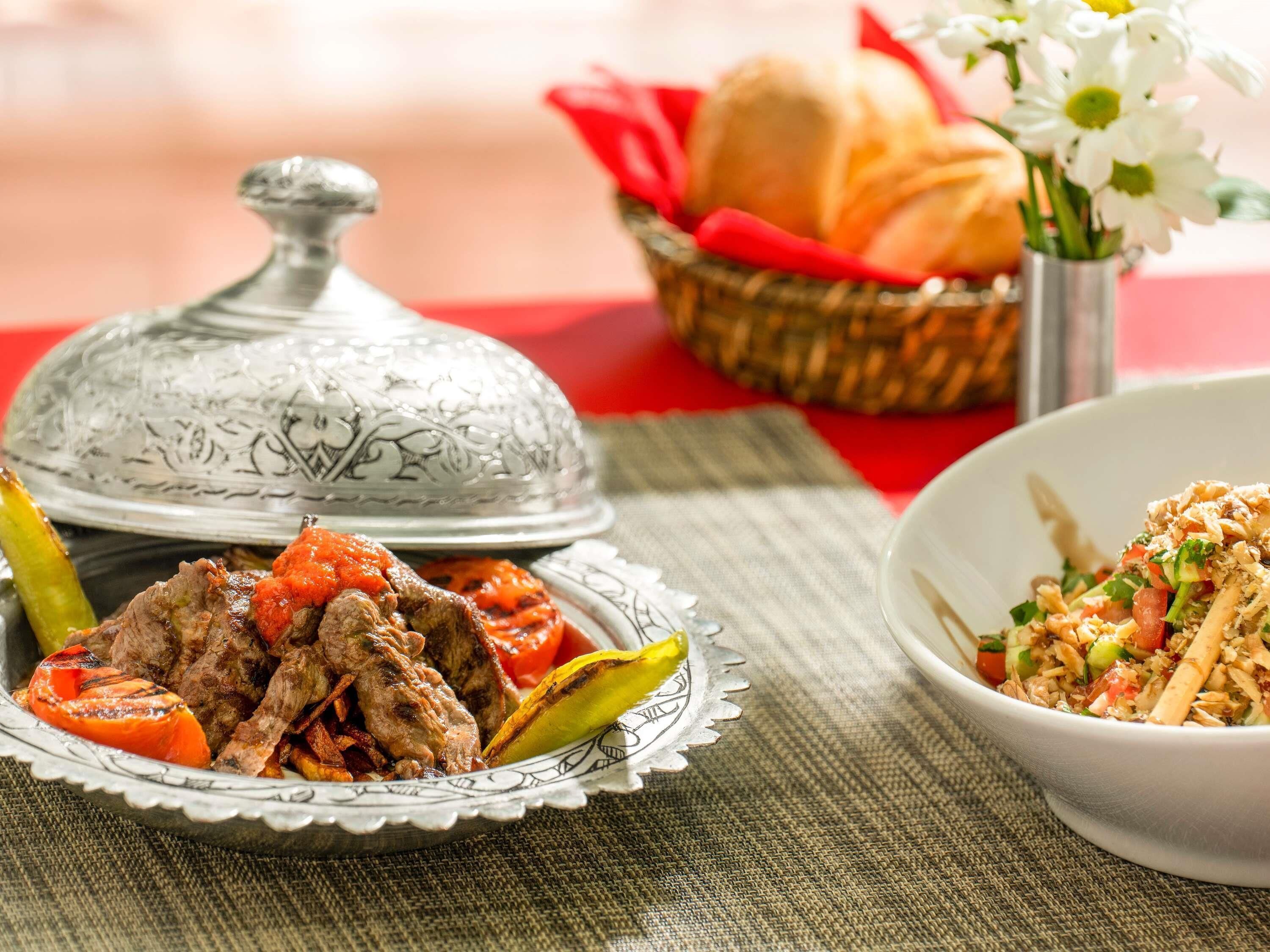 A dining table with a plate of grilled meat, a bowl of salad, a basket with bread, and a decorative silver cover.