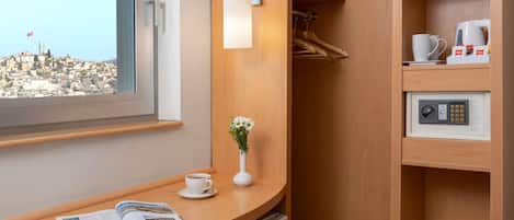 A hotel room with a wooden desk, a coffee machine, a minibar, and a view of a cityscape.