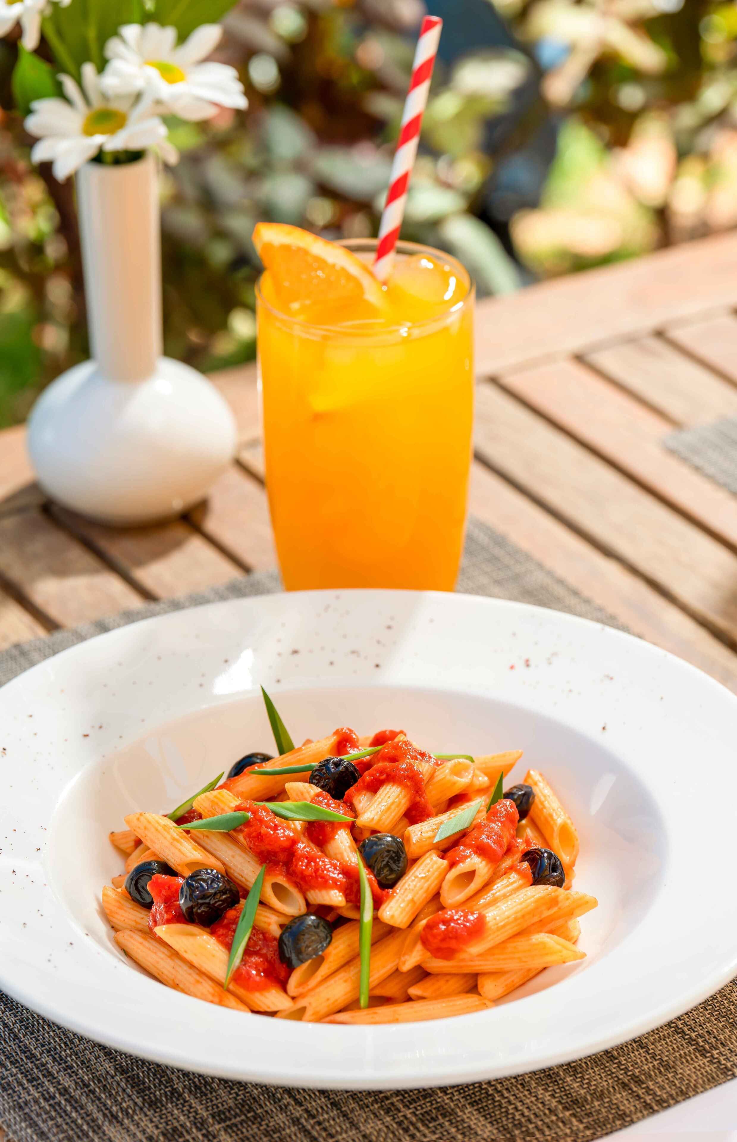 A plate of pasta with a red sauce and black olives, a glass of orange juice with a striped straw, and a white vase with flowers in the background.