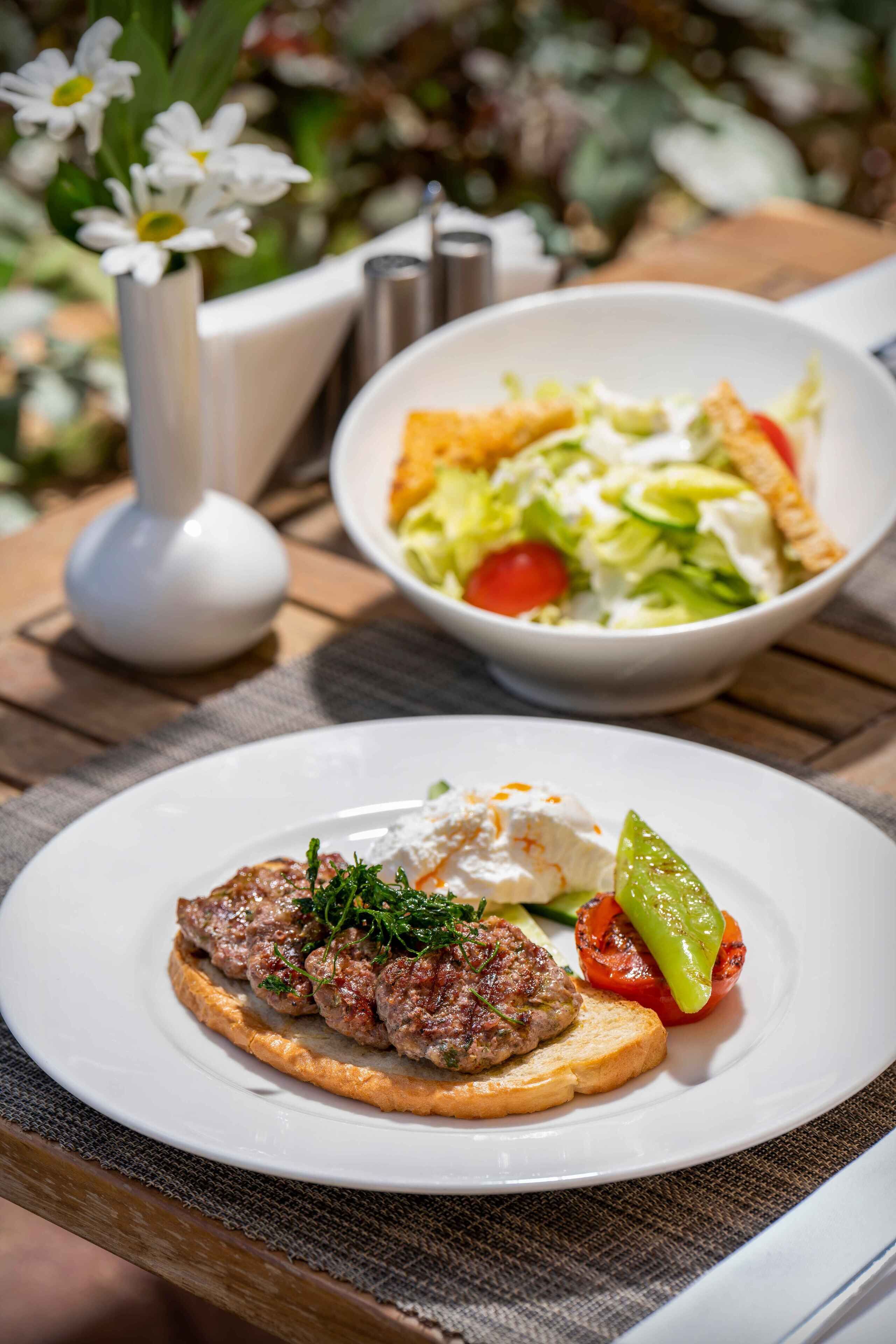 A plate of food with a meat patty, a slice of bread, and a grilled vegetable, accompanied by a bowl of salad.