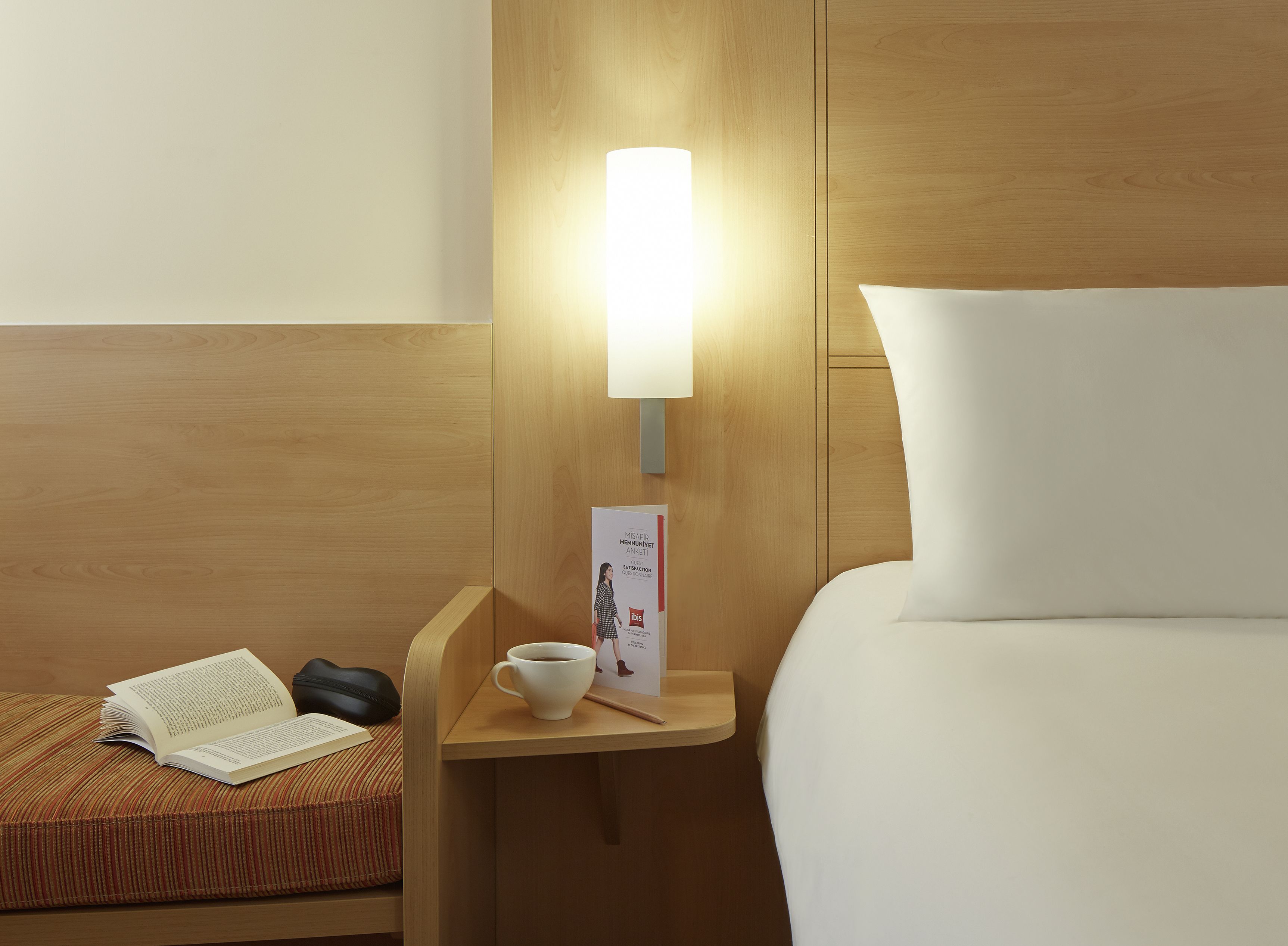 A hotel room with a neatly made bed, a bedside table with a lamp, a cup of coffee, and a book.