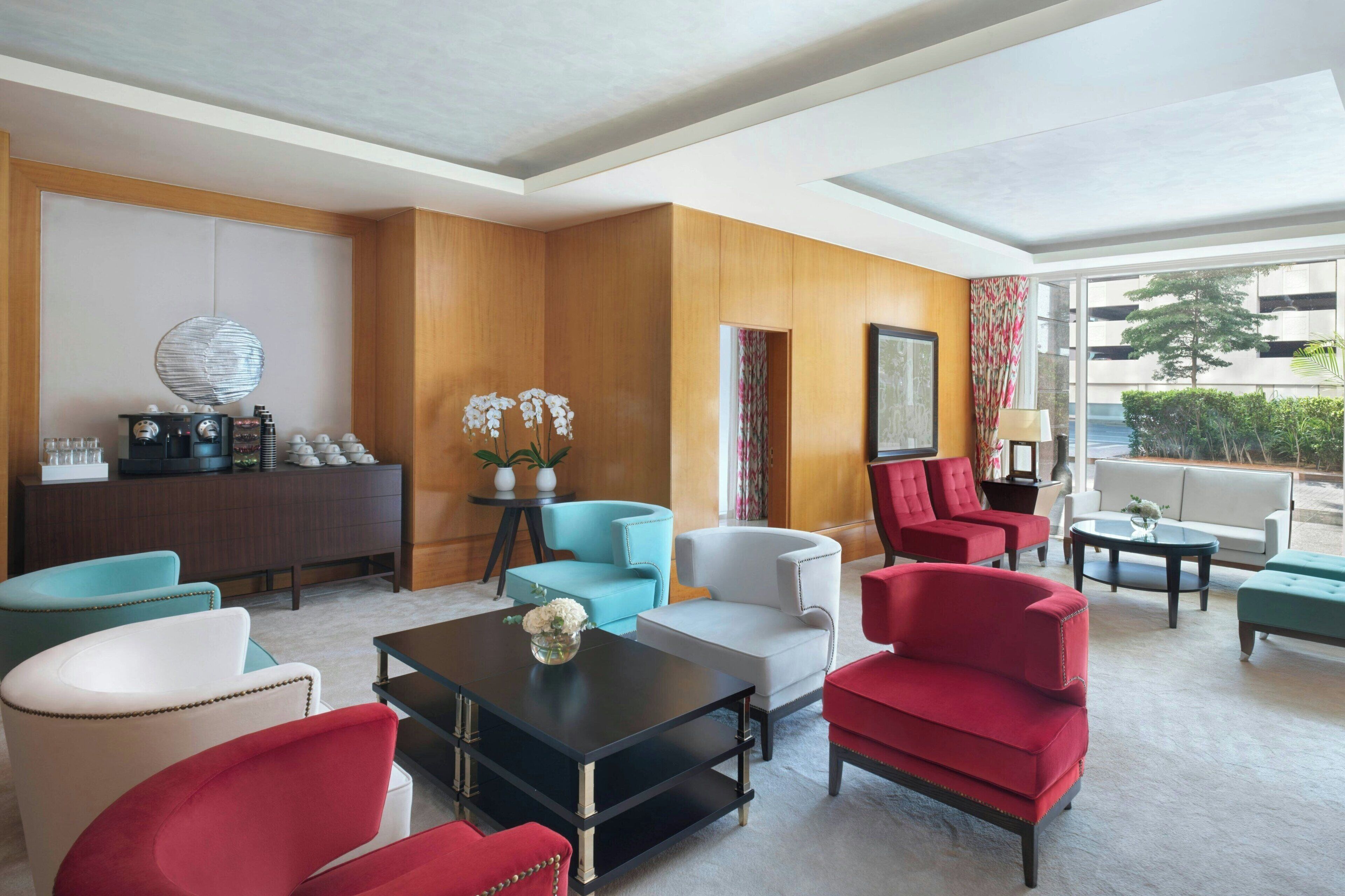 A hotel lobby with a wooden panel wall, a coffee machine, and various seating options including red and white armchairs and a sofa.