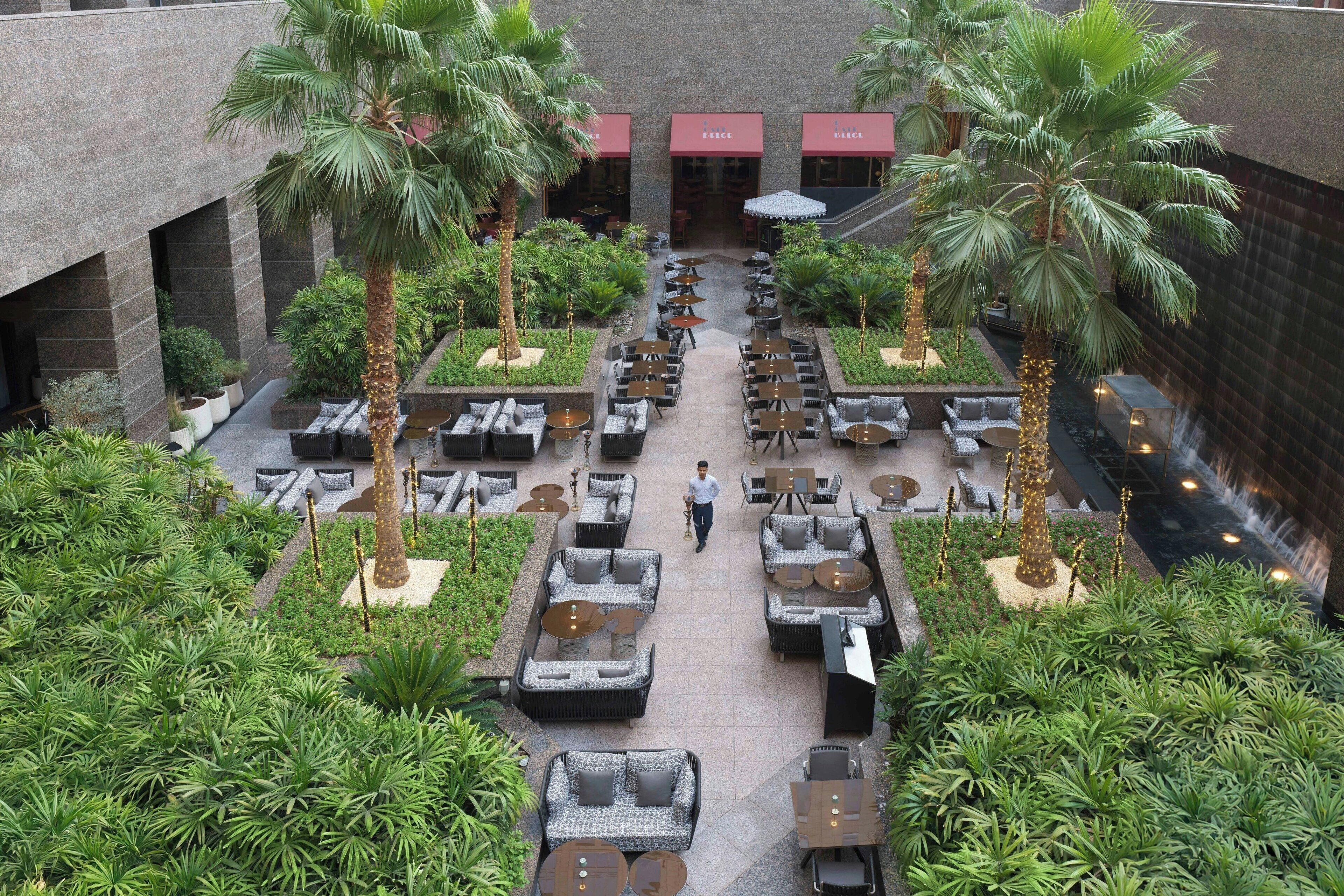 An outdoor dining area with multiple tables and chairs, surrounded by palm trees and greenery.