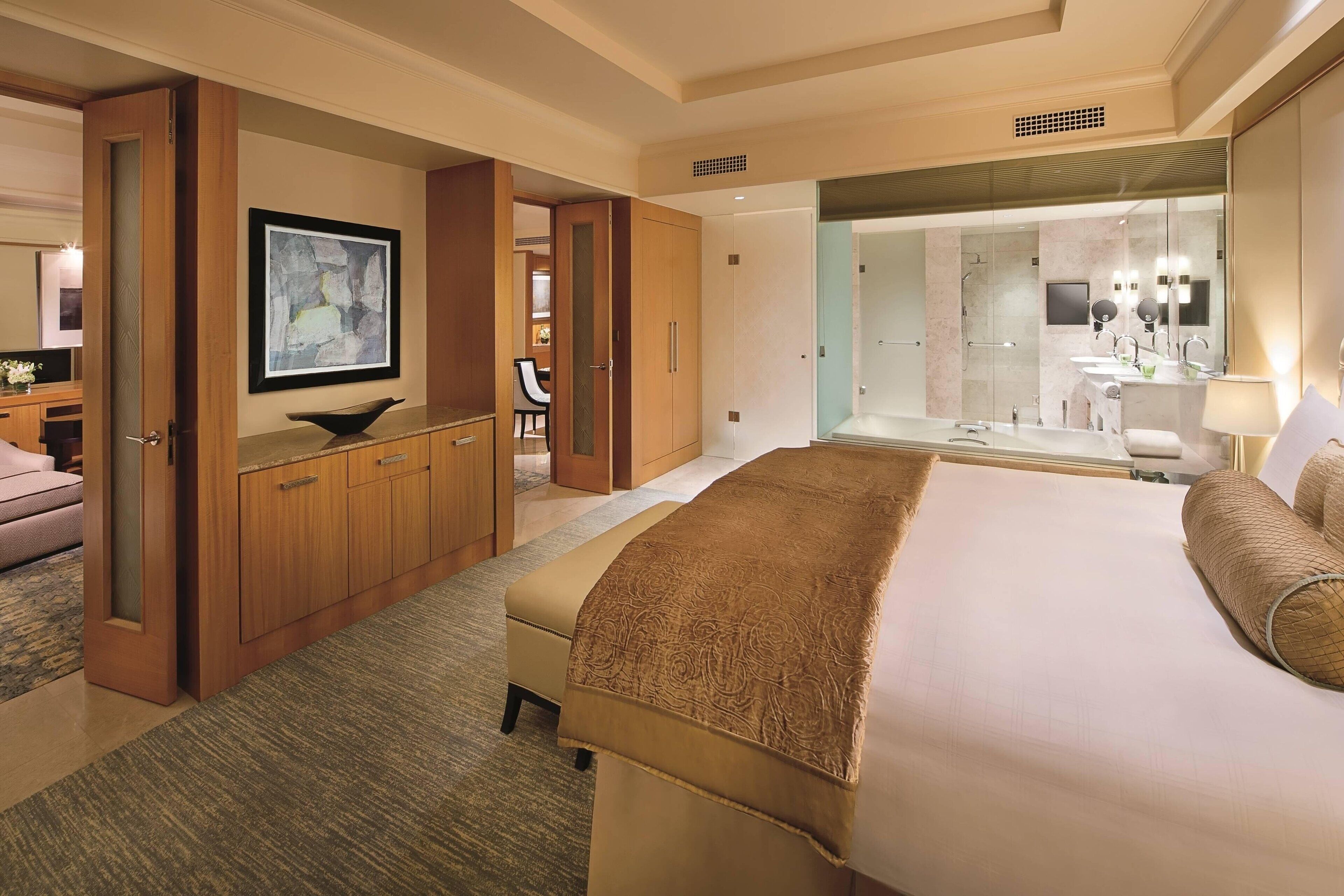 A hotel room with a large bed, a wooden wardrobe, a desk with a chair, a bathroom with a glass shower, and a framed abstract painting on the wall.