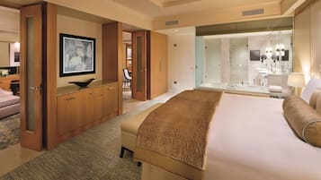 A hotel room with a large bed, a wooden wardrobe, a desk with a chair, a bathroom with a glass shower, and a framed abstract painting on the wall.