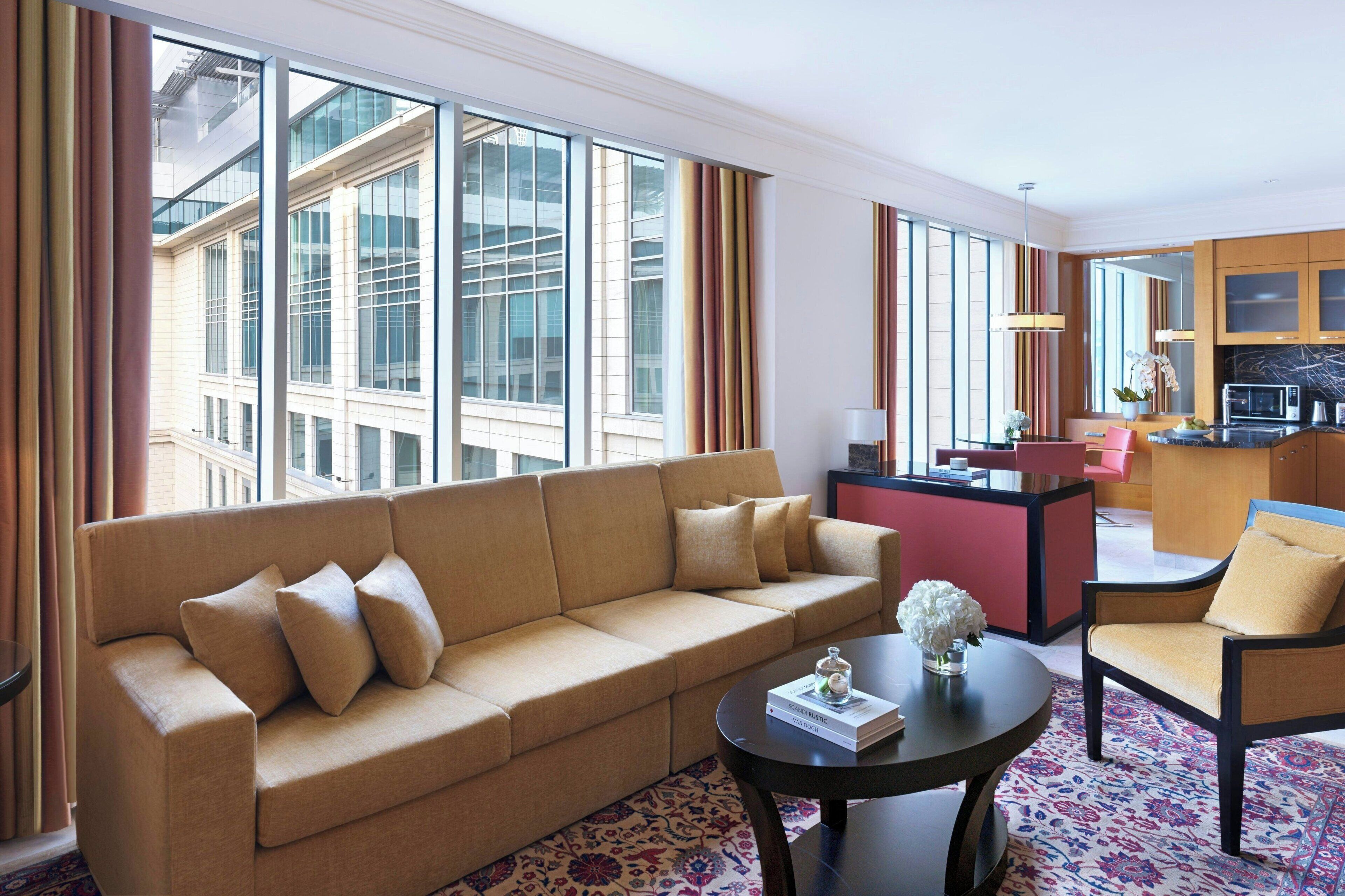 A modern hotel room with a beige sofa, a round coffee table, and a small kitchen area.