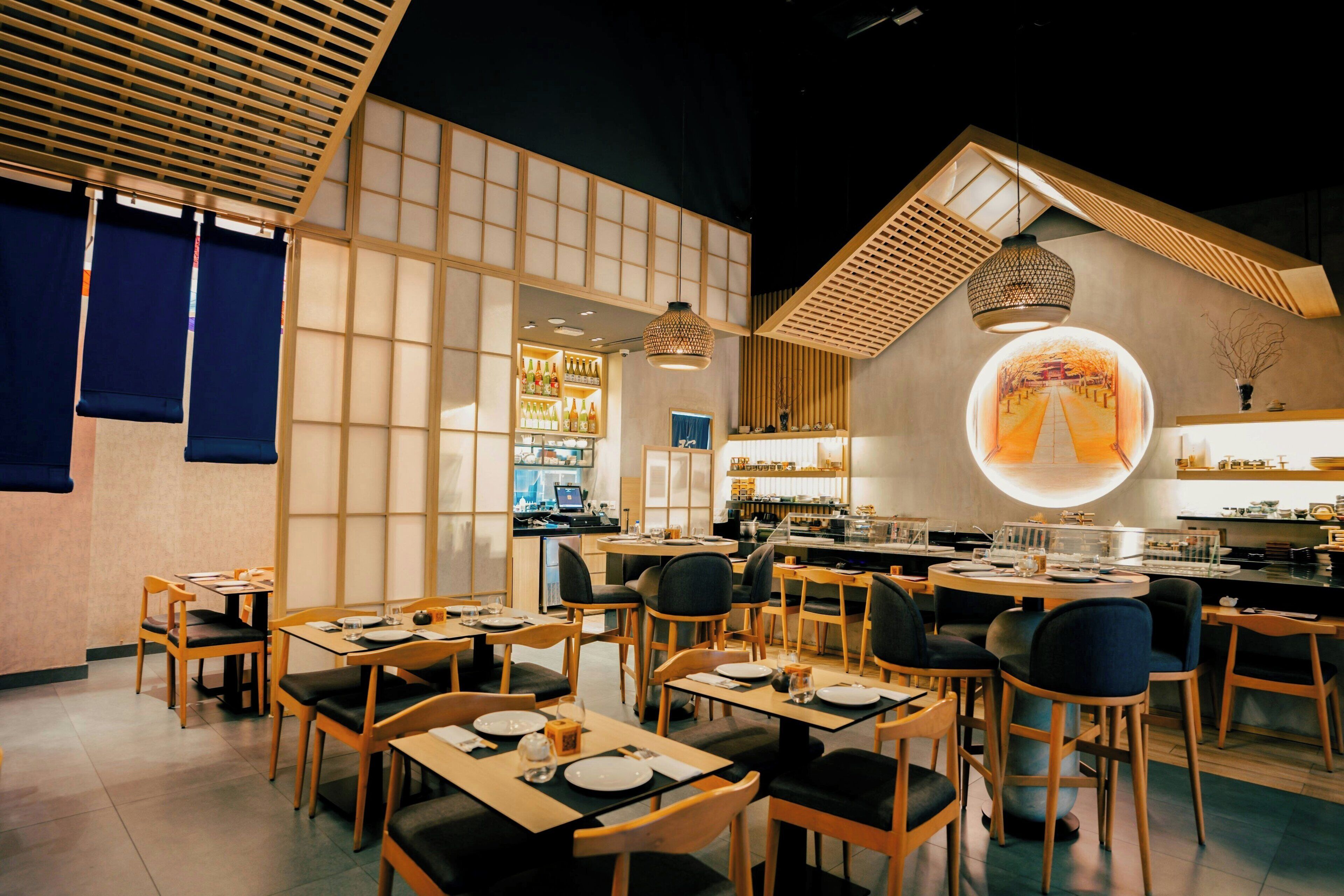 L’intérieur d’un restaurant, avec des meubles en bois, un espace de restauration central et une cuisine visible au fond.