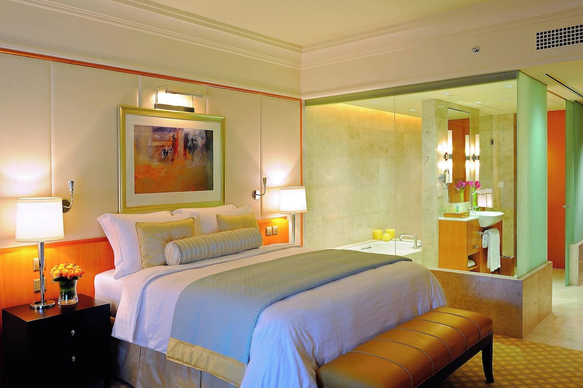 Quarto de hotel com uma cama grande, luminárias de cabeceira, mesa de cabeceira com um vaso de flores e um banheiro visível através de uma porta de vidro.