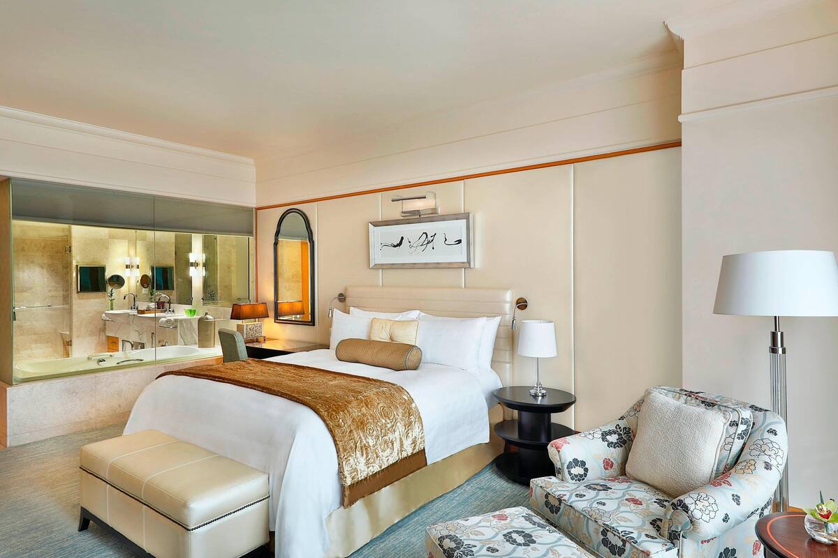 Quarto de hotel com uma cama grande, um espaço de estar com um sofá floral, uma pequena mesa redonda e um espelho refletindo o quarto.