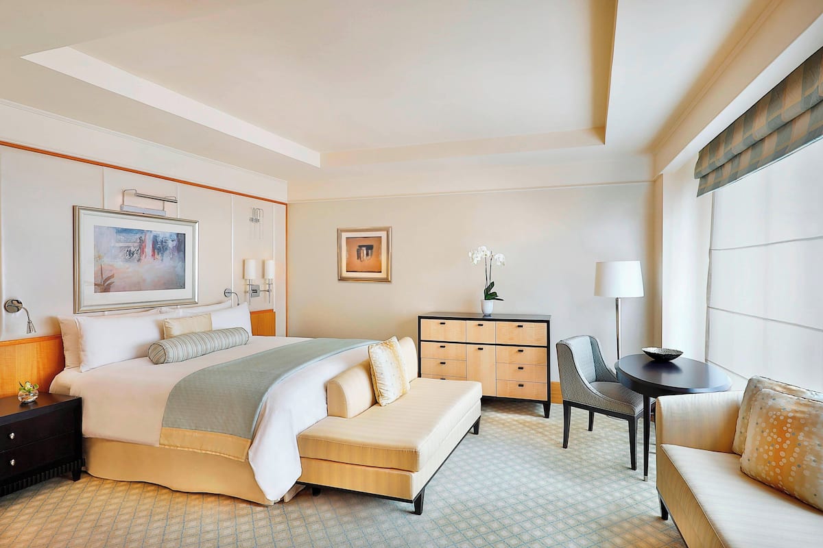 Quarto de hotel com uma cama grande, penteadeira, cadeira, mesa pequena e abajur.