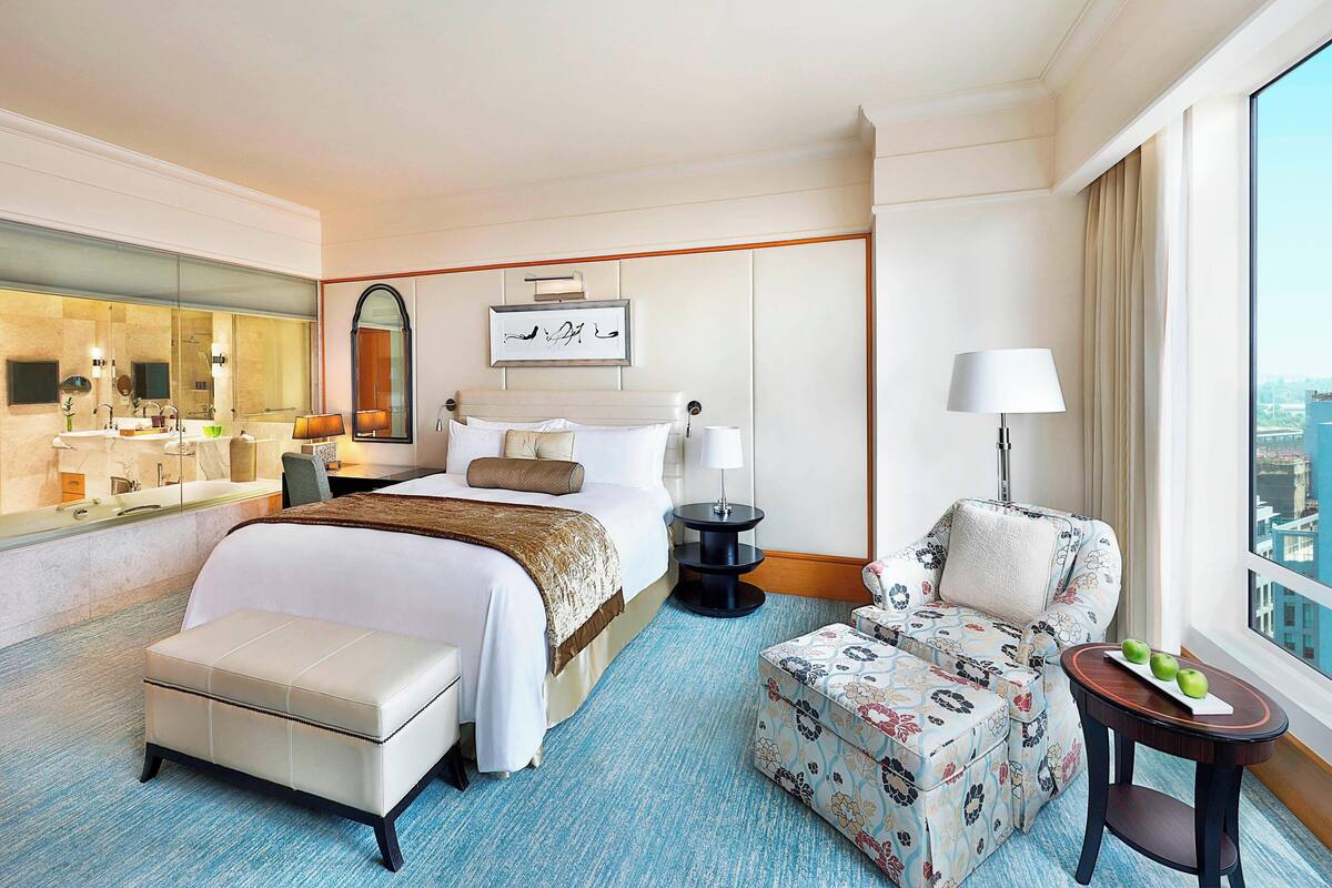 Quarto de hotel com uma cama grande, um espaço de estar com uma poltrona e um banquinho, uma pequena mesa com maçãs, um espelho e uma janela com vista para a cidade.