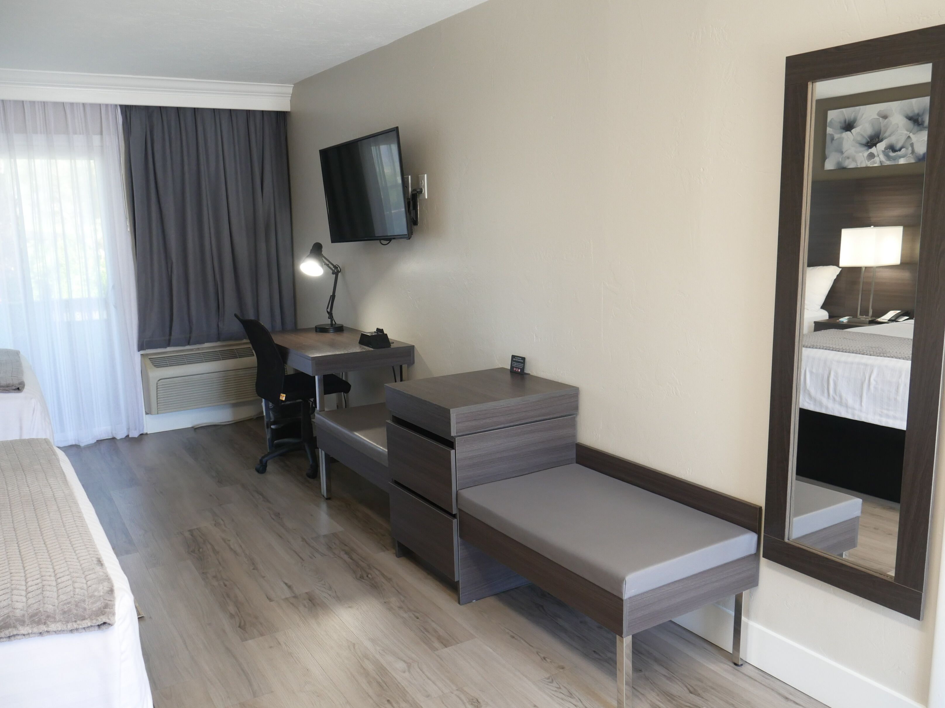 Hotellihuone, jossa on sänky, työpöytä tuolilla, työpöytälamppu, taulutelevisio ja suuri peili.