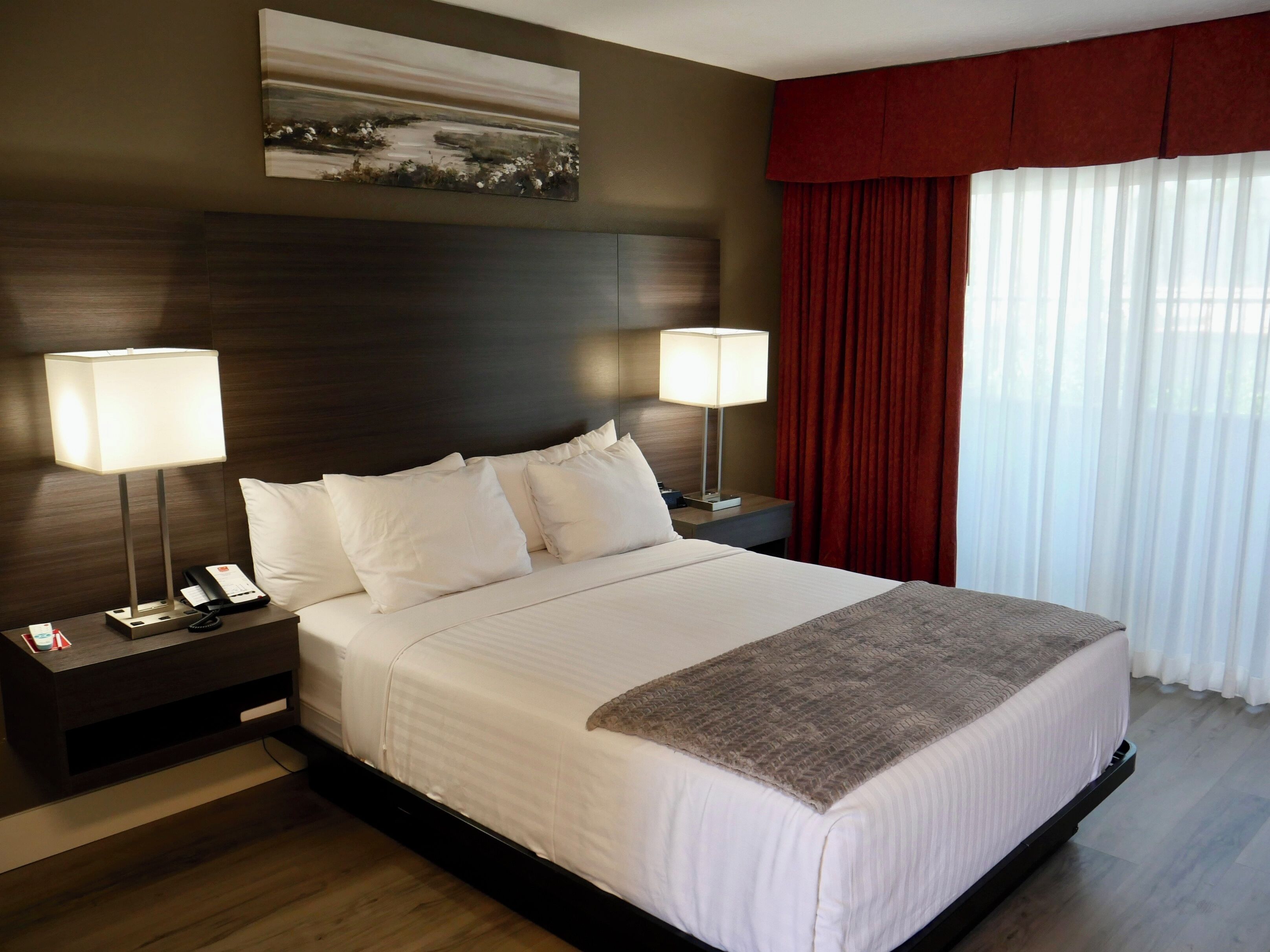A hotel room with a large bed, two bedside tables with lamps, a painting on the wall, and a window with curtains.