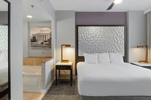 Une chambre d’hôtel avec un grand lit, une table de chevet, une lampe, une salle de bain avec baignoire et une œuvre d’art accrochée au mur.