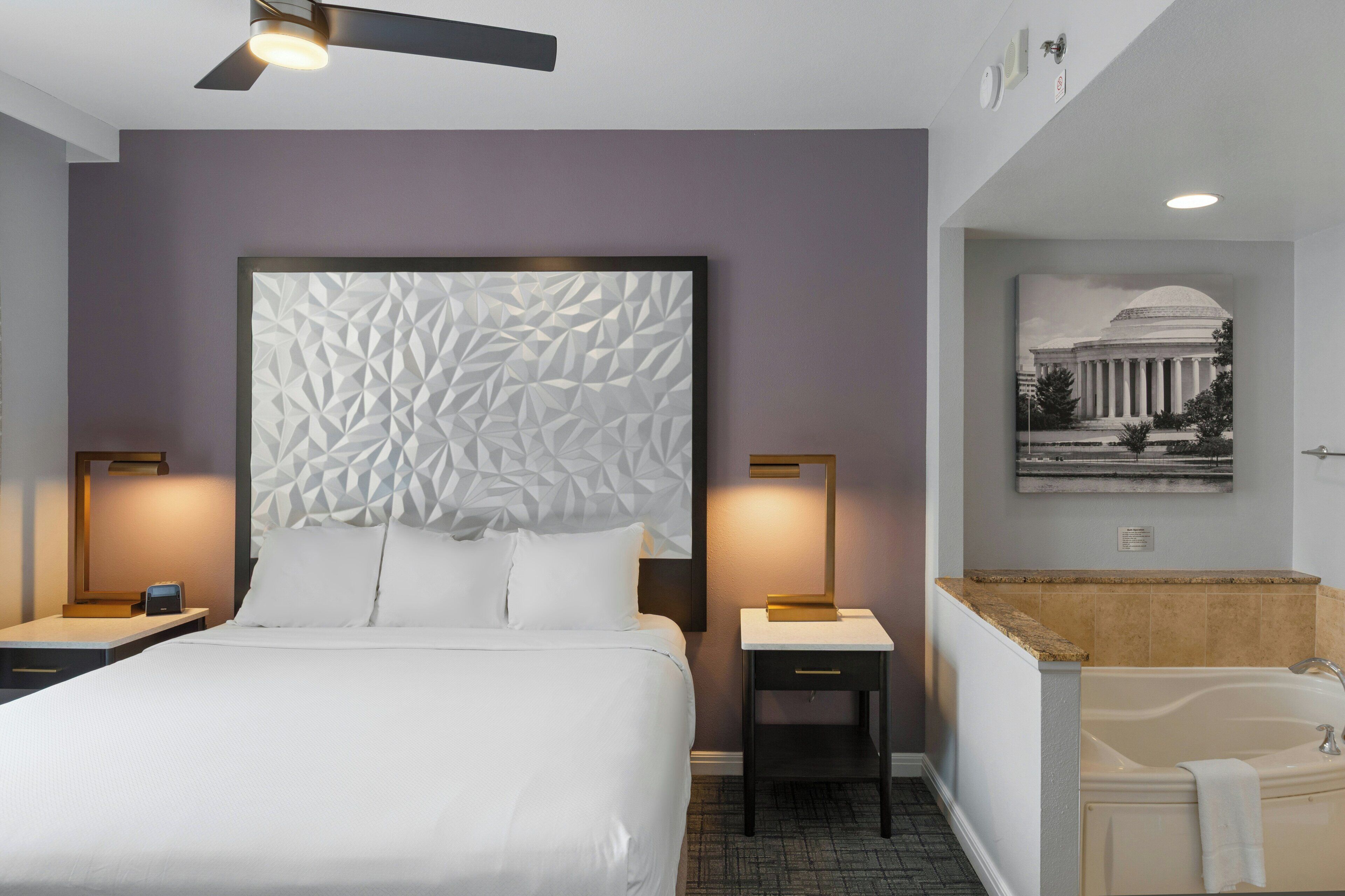 A hotel room with a bed, nightstands, a desk, a wall-mounted lamp, a painting, and a bathtub.