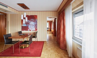 Quarto de hotel moderno com mesa de jantar de madeira, cadeiras pretas, tapete vermelho e uma grande janela com cortinas.