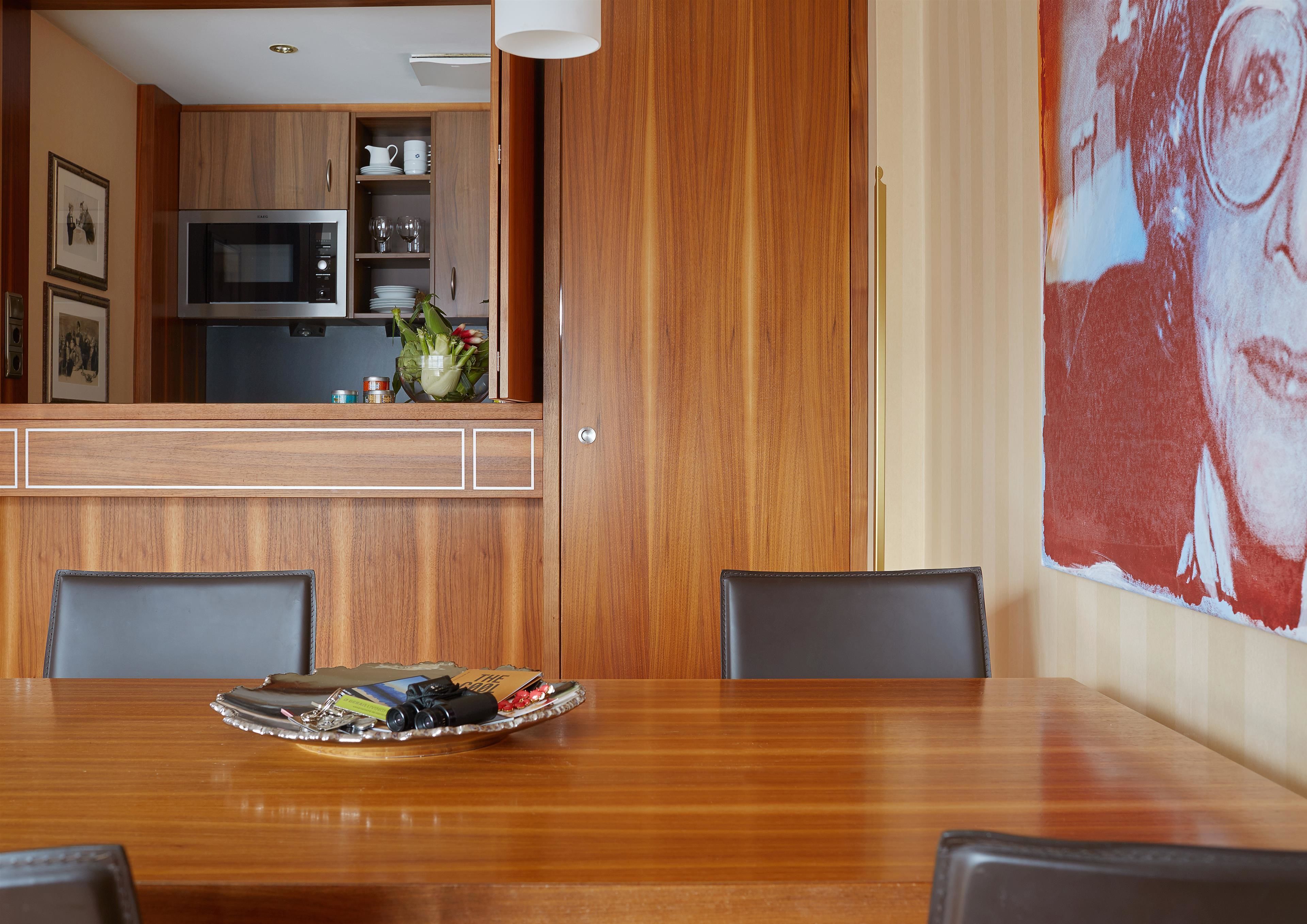 A wooden dining table with chairs, a kitchen area with wooden cabinets, and a large painting on the wall.