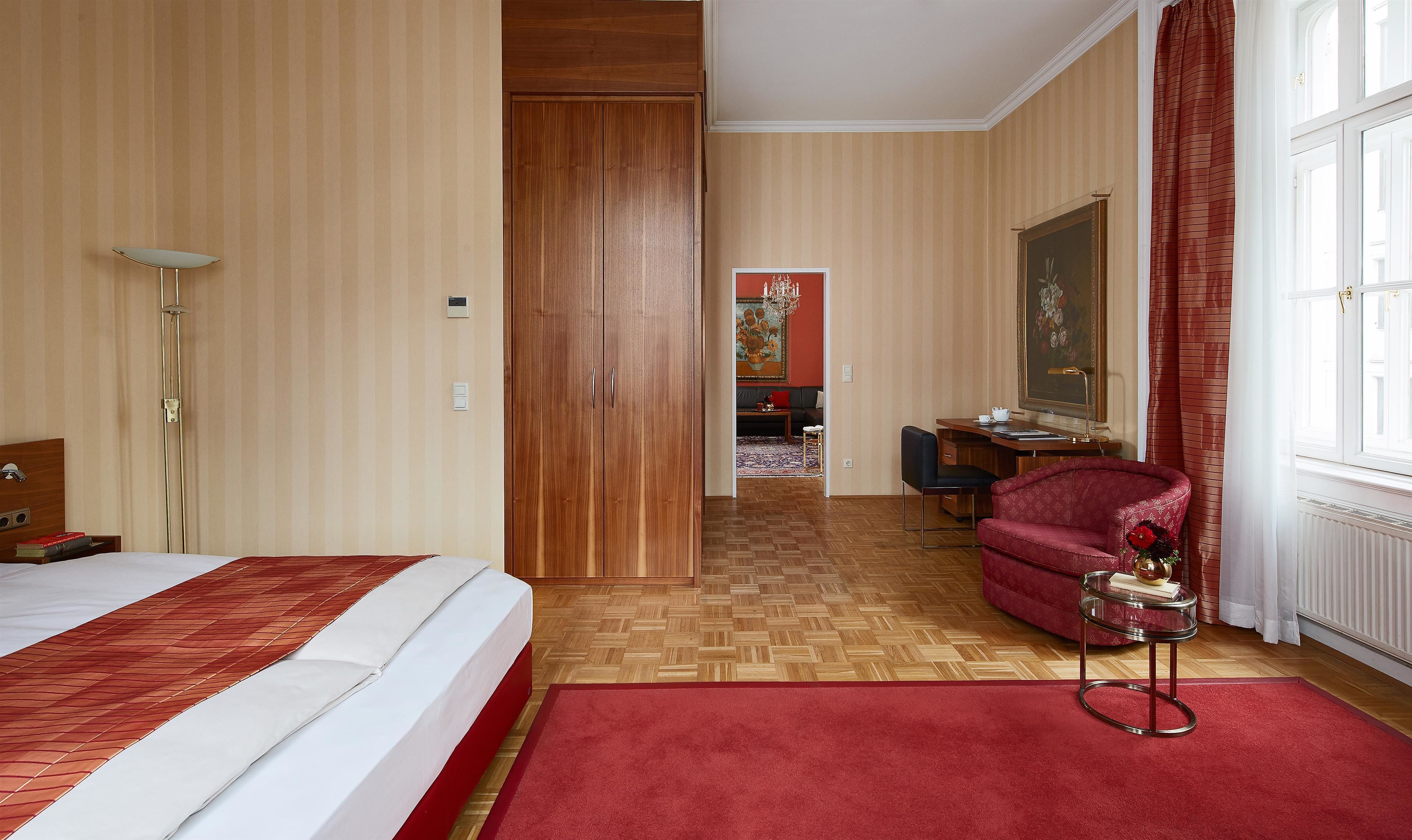 Une chambre d’hôtel avec un lit, un bureau, une chaise, un fauteuil rouge et une grande fenêtre avec des rideaux.
