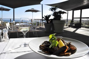 Una mesa de comedor con un plato de mejillones y una copa de vino blanco, frente a un fondo de sombrillas y plantas en macetas, con vistas a un paisaje pintoresco.
