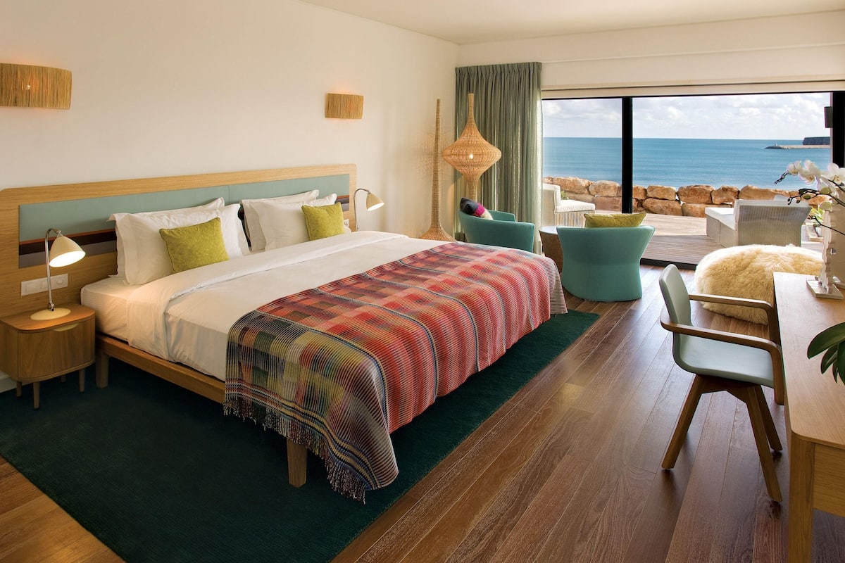 A hotel room with a large bed, a view of the ocean, a dining area, and a seating area with a view.
