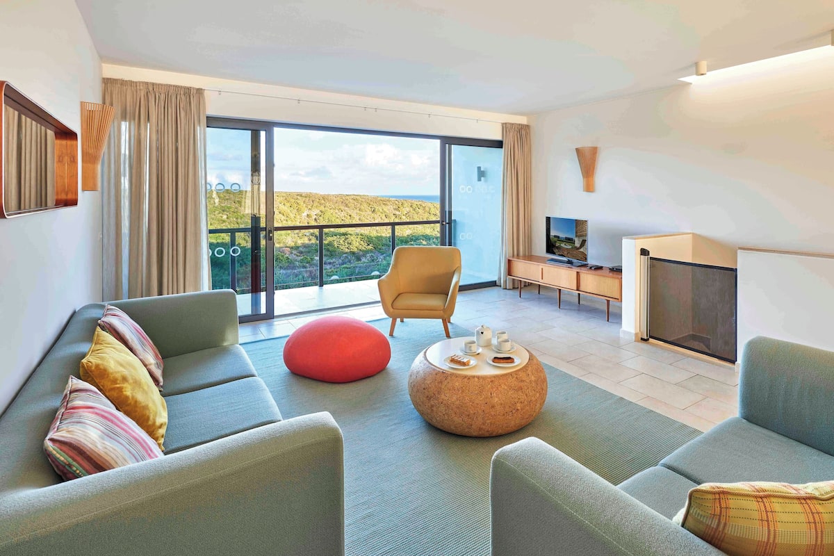 A modern living room with a sofa, armchairs, a red ottoman, and a round coffee table. There is a television and a fireplace. The room has large windows with a view of a mountainous landscape.