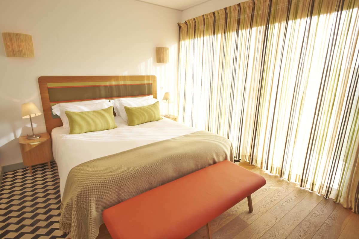 A hotel room with a large bed, two bedside tables, a bench, and a window with curtains.