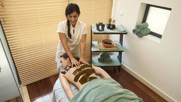 A massage therapy room with a wooden floor, a massage table, a massage therapist, and a small table with towels and a bowl.