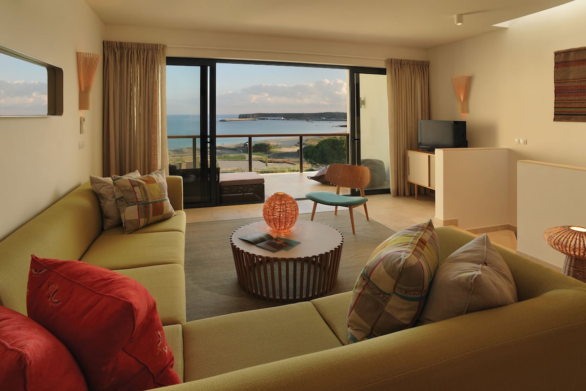 A living room with a sofa, a coffee table, a television, and a view of the sea.