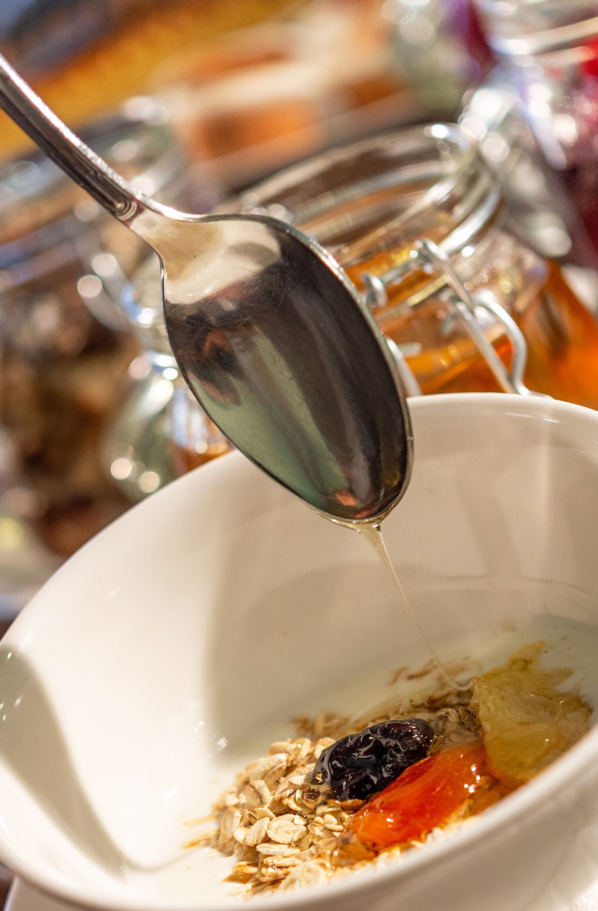 A spoon pouring a liquid into a bowl containing oats, a raisin, and a piece of fruit.