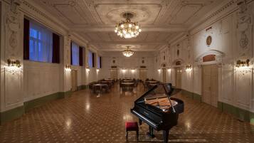 A grand hall with a grand piano, ornate ceiling, and rows of tables and chairs.