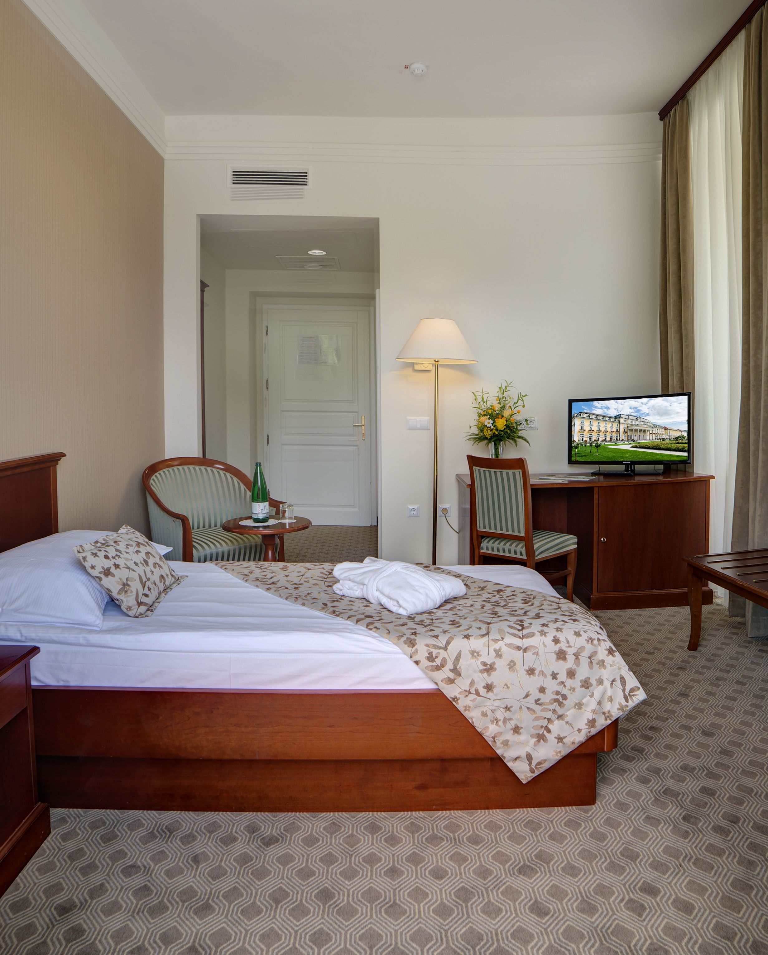 A hotel room with a large bed, a desk with a television, a chair, and a lamp.
