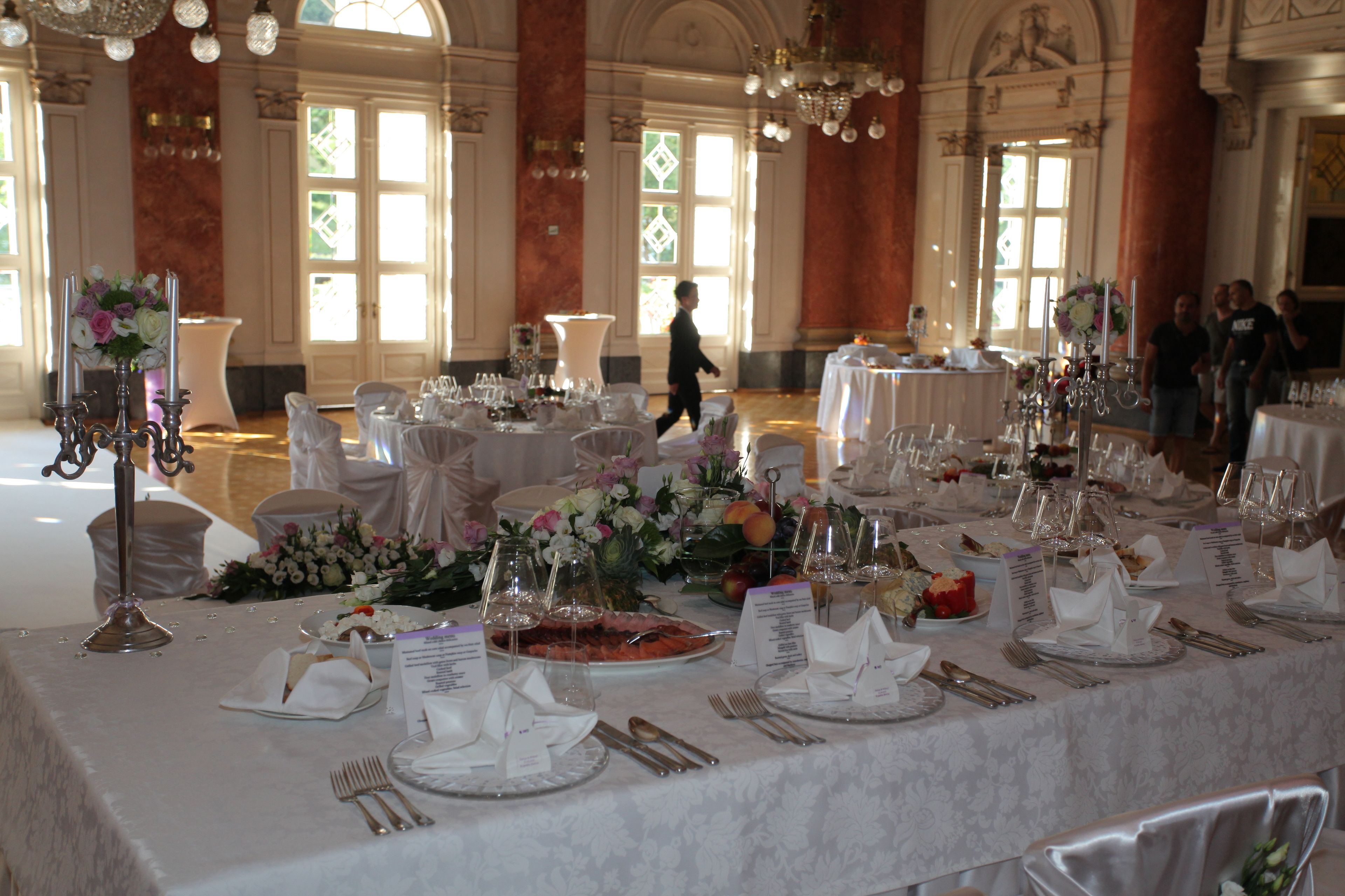 A banquet hall with round tables set for a formal event, featuring white tablecloths, floral centerpieces, and silverware.