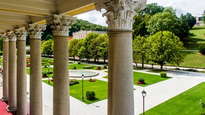 A covered walkway with classical columns, a well-manicured garden with a fountain, and a building in the background.