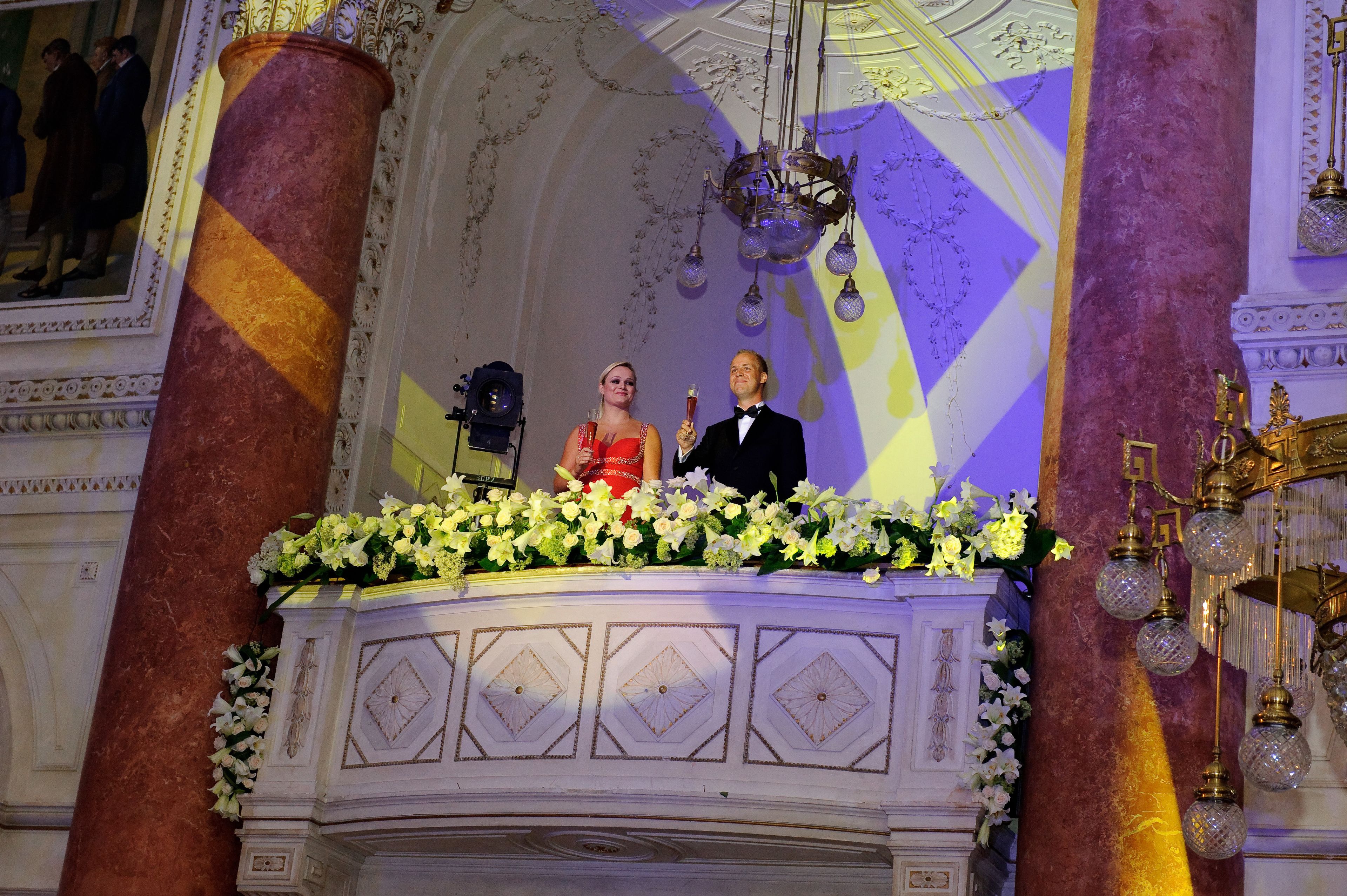 Un salón imponente con columnas ornamentadas, una araña y un arreglo floral sobre un atril.