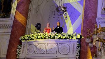Un salón imponente con columnas ornamentadas, una araña y un arreglo floral sobre un atril.