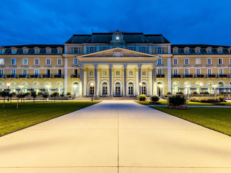 A large, symmetrical building with a central entrance flanked by columns, surrounded by a well-maintained lawn and a paved walkway.