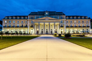 Un edificio grande y simétrico con una entrada central flanqueada por columnas, rodeado de un césped cuidado y un camino pavimentado.
