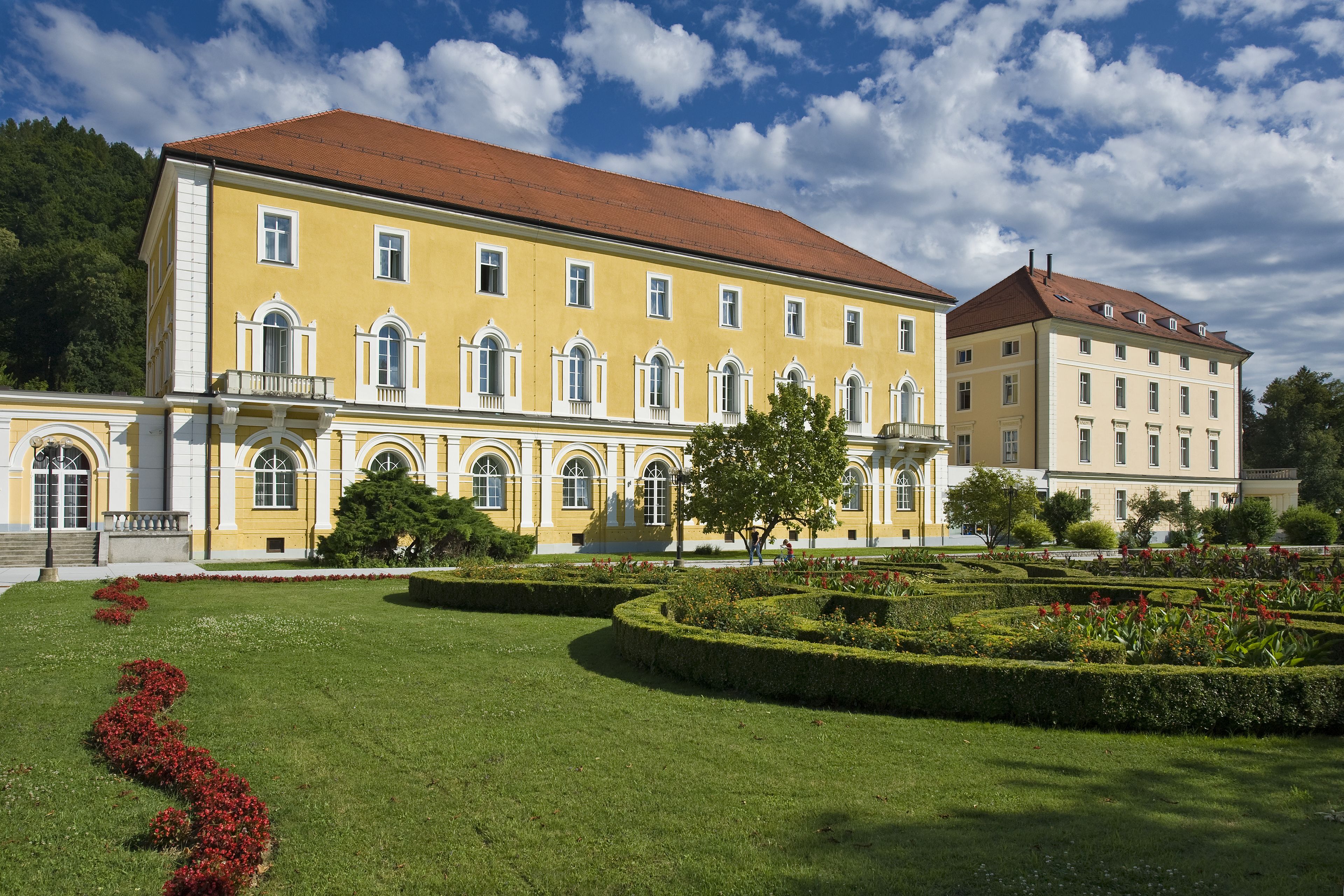 A large yellow building with a red roof, surrounded by well-maintained gardens and a clear sky.