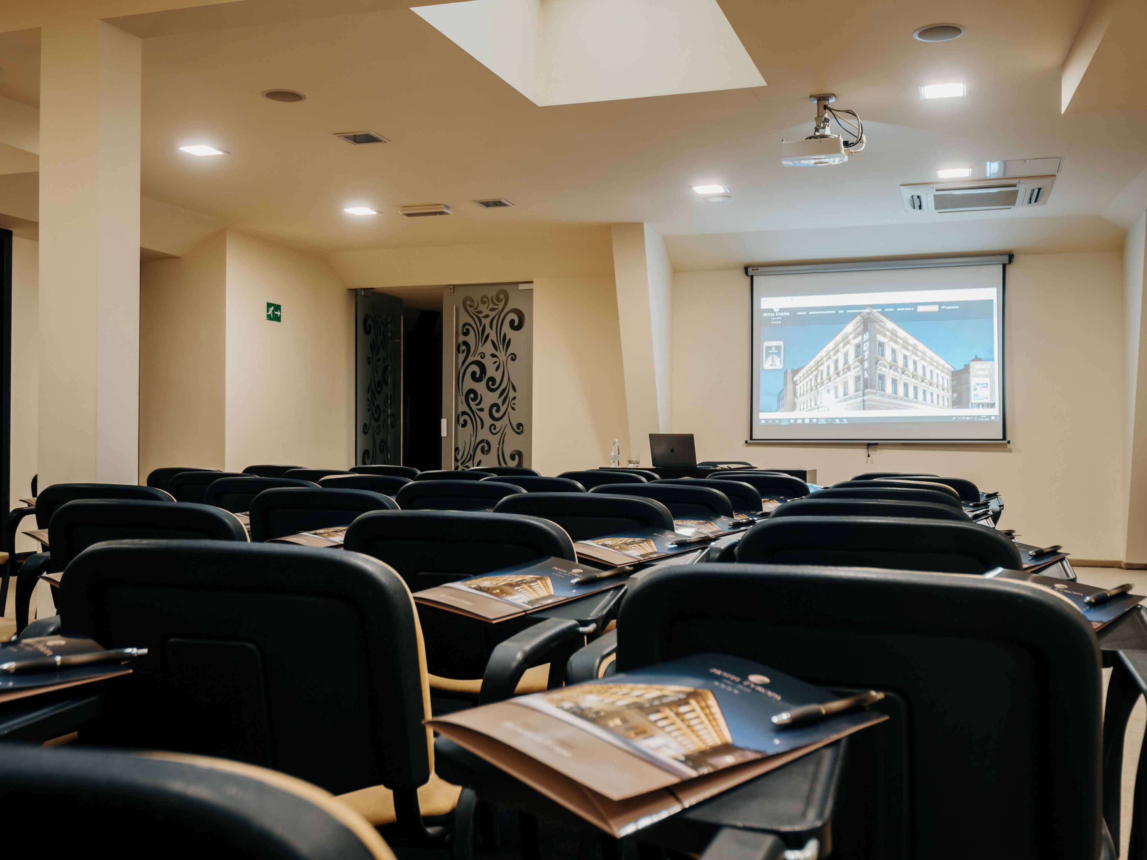 A conference room with rows of black chairs, a projector screen displaying a building image, and a laptop on a table.