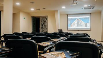 Une salle de conférence avec des rangées de chaises noires, un écran de projection affichant l’image d’un bâtiment et un ordinateur portable posé sur une table.