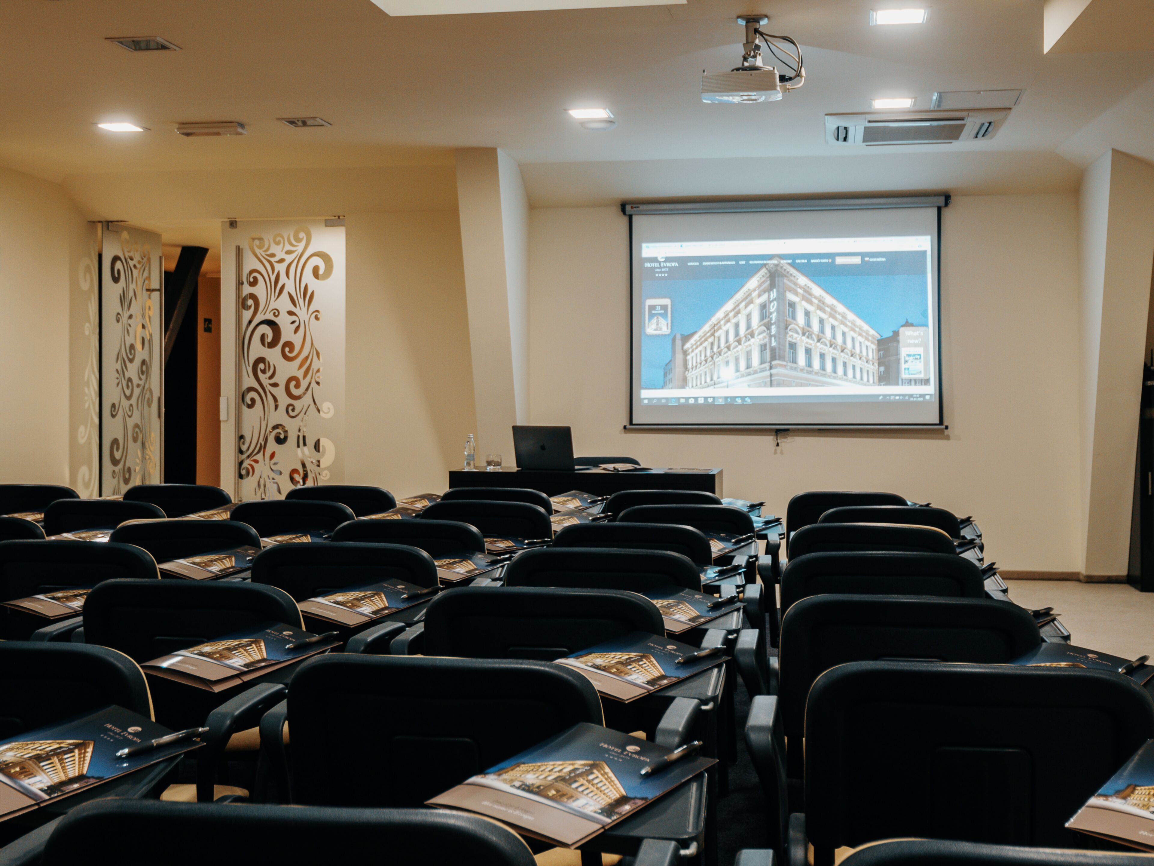 Un salón de conferencias con filas de sillas, una pantalla de proyector mostrando una imagen de un edificio y un portátil sobre una mesa.