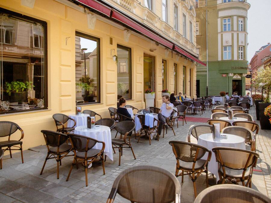An outdoor cafe with tables and chairs, large windows, and a building with a green facade.