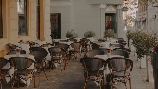 Área de refeições ao ar livre com cadeiras de vime e toalhas de mesa brancas, em frente a uma fachada de prédio com janelas amplas e uma porta.