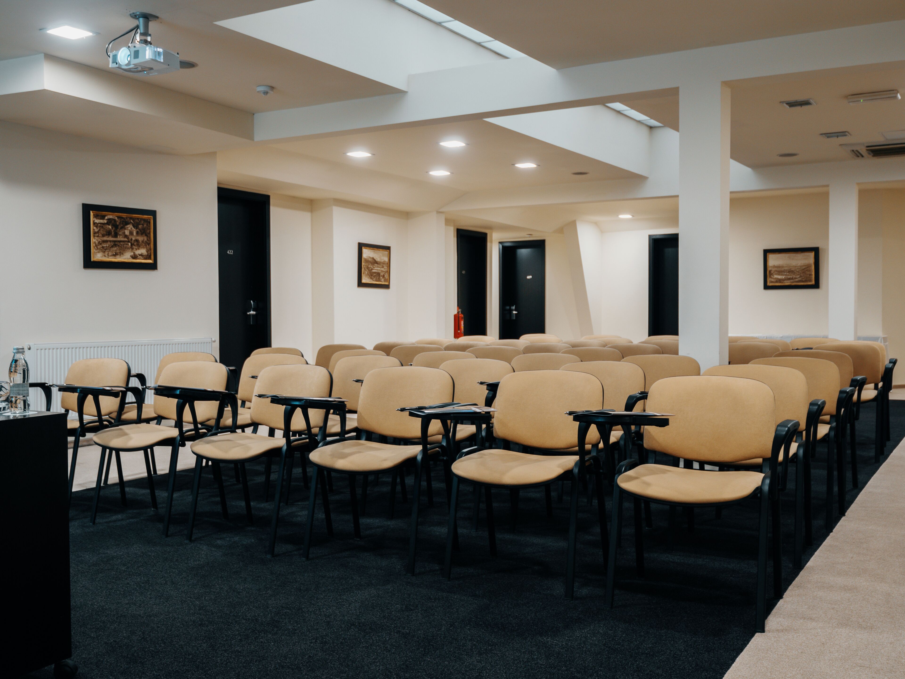 Une salle de conférence avec des rangées de chaises beiges, des tables noires et des luminaires au plafond.
